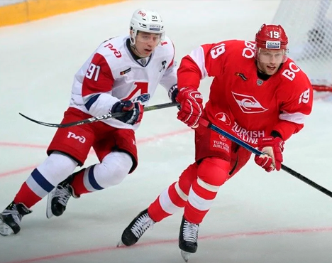 Двое хоккеистов в движении на льду. Игрок в красной форме ведет шайбу, а спортсмен в белой форме с синими и красными элементами пытается его догнать. Видны яркие логотипы на форме