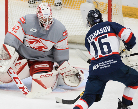 Хоккейный момент: вратарь в серо-красной форме защищает ворота от игрока в темно-синей форме с номером 16. Игрок готовится сделать бросок. На льду видны логотипы и экипировка