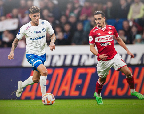 На изображении два футболиста в игровой момент. Игрок в бело-синей форме ведет мяч, а футболист в красной форме соперника старается пресечь его действия. На заднем плане видны трибуны с болельщиками